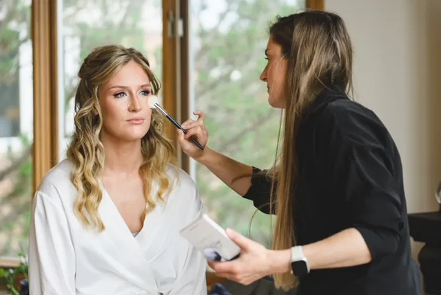 Bride in a white robe receiving soft glam bridal makeup before her wedding